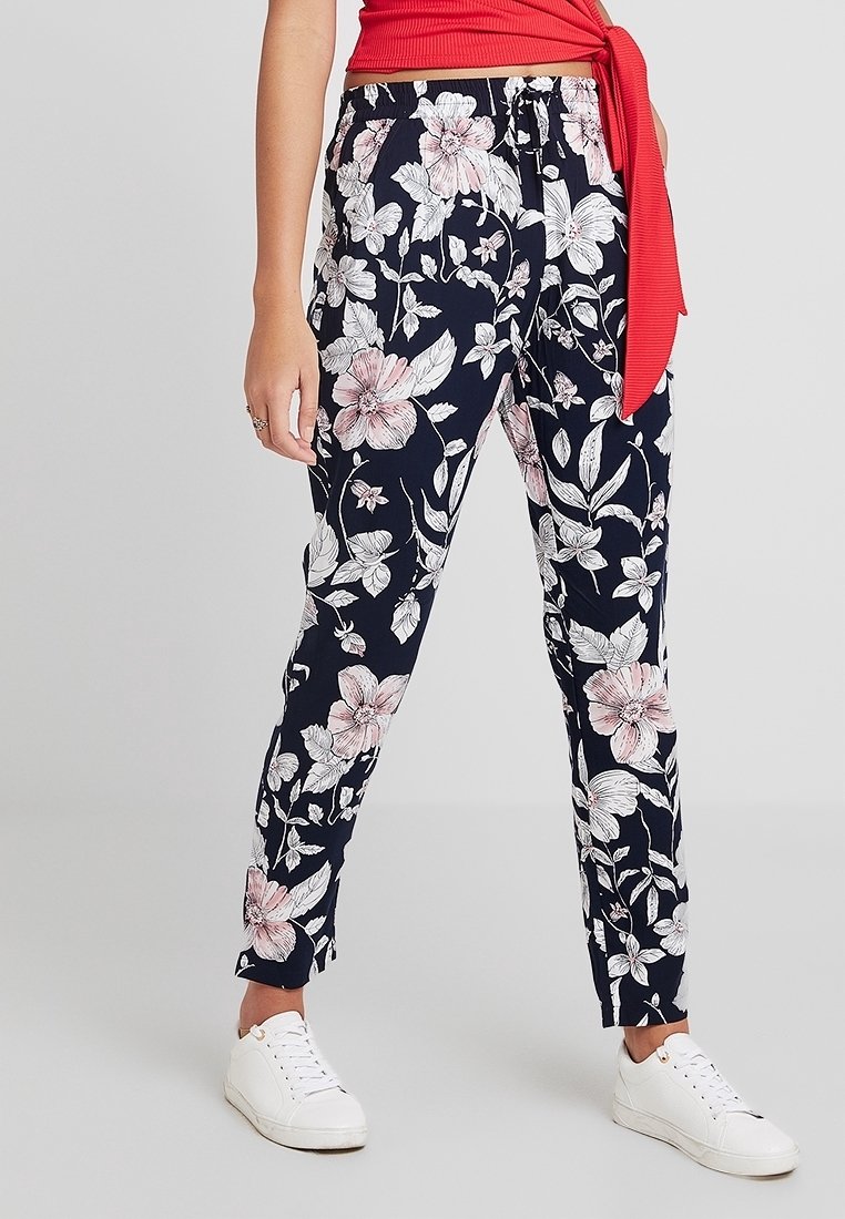 Woman wearing navy floral print pants, white sneakers, and a red cropped top with a side tie, standing against a plain background.