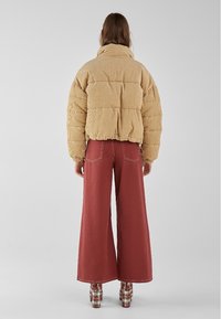 Chaqueta acolchada de pana color tan con cuello alto y textura abullonada; combinada con pantalones burdeos de pierna ancha y zapatos de cuadros.