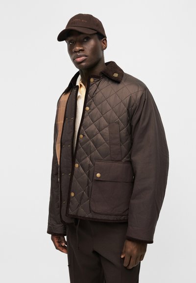 Moški v temno rjavi prešiti jakni z žepki na sprednji strani, temno rjavi kapici in svetli srajci, stoji pred enobarvnim svetlim ozadjem.