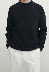 Personne portant un pull en tricot texturé noir à manches longues et un pantalon plissé blanc, les mains dans les poches, devant un fond neutre.