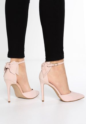 Femme portant des chaussures à talons hauts rose pâle avec des brides à la cheville et de grands nœuds, associées à un pantalon noir ajusté sur fond blanc.