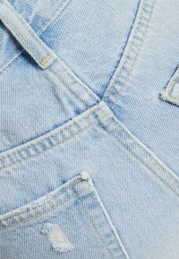 Ljusblå denimoveraller med strukturerad tyg, framträdande sömmar och två framfickor med fransade kanter och diagonala sömmar.