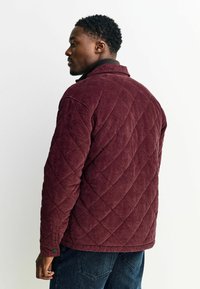 Bordeaux quilted corduroy jack met ruitpatroon, kraag en knoopmanchetten, gedragen over donkere jeans tegen een lichte achtergrond.