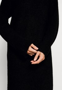 Svart stickad tröja med hög krage och långa ärmar. Tyget ser mjukt och strukturerat ut, med breda muddar och en avslappnad passform.