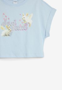 Lyseblå cropped t-shirt med korte ærmer, der præsenterer illustrerede hvide kaniner med vinger og glitrende "desigual" tekst i pink og sølv.