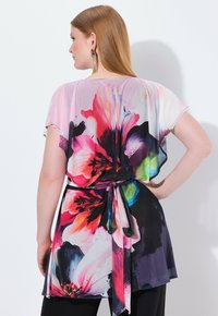 Femme aux longs cheveux roux portant une tunique florale colorée avec une ceinture à nouer, vue de dos sur un fond blanc uni.