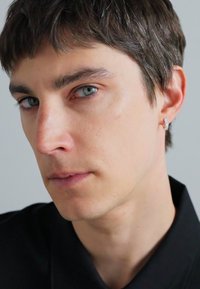 Portret osobe s kratkom smeškom kosom i plavim očima, koja nosi crnu košulju s ovratnikom i sjajnu srebrnu naušnicu s ukrasnim dizajnom.