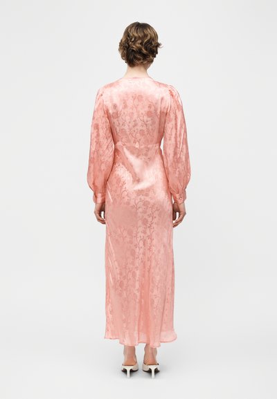 Γυναίκα με κοντά σγουρά μαλλιά που φοράει ένα μακρύ ροζ μεταξωτό φόρεμα με ανθικό μοτίβο και φουσκωτά μακριά μανίκια, στέκεται με την πλάτη προς τα εμπρός.