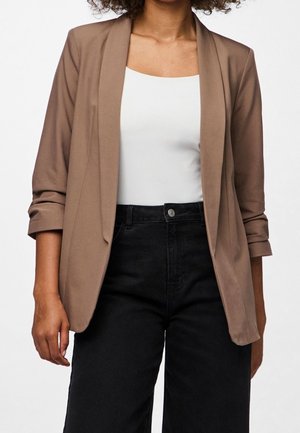 Personne portant un blazer marron clair aux manches retroussées, un haut blanc et un pantalon noir taille haute sur un fond clair uni.