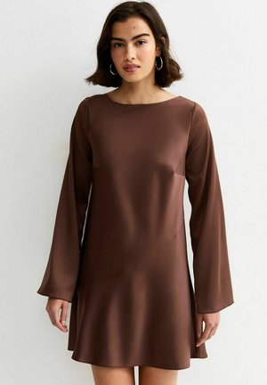 Vrouw met halflang donker haar, gekleed in een bruine satijnen mini-jurk met lange mouwen en wijduitlopende mouwen, en met ronde oorbellen, staand tegen een witte muur.