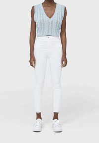 Top de punto sin mangas en azul claro con escote en V, combinado con jeans blancos ajustados. Las zapatillas son totalmente blancas. En general, un atuendo casual moderno.