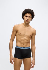Cuecas boxer pretas com cós azul claro com marca, apresentando textura suave e design ajustado. O modelo está sem camisa, exibindo um físico definido.