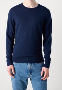Maglione lavorato a maglia blu navy con scollo rotondo e maniche lunghe, caratterizzato da polsini e orlo a coste, abbinato a jeans di denim blu chiaro.