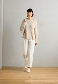 Pull beige à côtes avec des manches longues, associé à un pantalon large blanc et des baskets blanches. Le mannequin porte un petit sac beige.