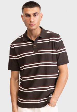 Νεαρός άνδρας με κοντά μαλλιά, φοράει καφέ πλεκτό πόλο με κοντά μανίκια και οριζόντιες λευκές και κόκκινες ρίγες, με το χέρι στην τσέπη, ουδέτερο φόντο.