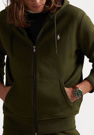 Ανδρική ζακέτα φούτερ σε ελαιόλαδο με φερμουάρ, μπροστινές τσέπες, ριμπ μανσέτες και μικρό λευκό λογότυπο. Ένα καφέ ρολόι με πράσινο καντράν φοριέται στον καρπό.