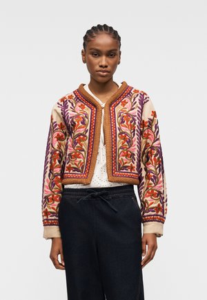 Jeune femme portant une veste courte crème brodée de fleurs colorées violettes, roses et oranges, sur une blouse blanche et un pantalon foncé.