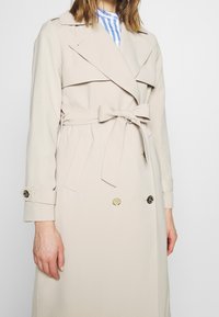 Femme portant un trench-coat beige ceinturé avec des boutons croisés et des lanières aux poignets sur une chemise rayée bleue et blanche.