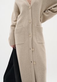 Beige stickad cardigan med v-ringning, stora framfickor och sex framknappar. Har ribbade mudd och en mjuk textur.