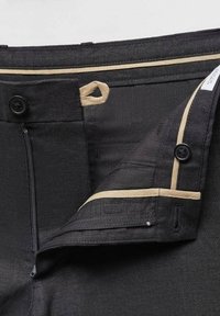 Pantalons noirs sur mesure avec des finitions beiges, partiellement zippés, montrant les détails de bouton et de fermeture à agrafe le long de la taille.
