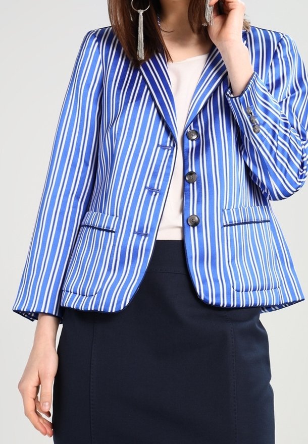 Femme portant un blazer bleu et blanc rayé sur un haut blanc et une jupe bleu marine foncé, avec de longues boucles d'oreilles à pampilles.