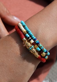Bracelet multi-brins avec des perles turquoises, rouges et dorées ; présente diverses textures et formes rondes ; superposé sur un poignet de teinte naturelle.