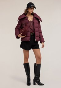 Giubbotto impermeabile bordeaux con ampio colletto, pantaloni corti neri a vita alta e stivali neri al ginocchio, che tiene una borsa bordeaux.