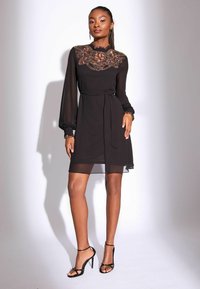 Melna caurspīdīga kleita ar izšūtu augstu kakla izgriezumu, garām piedurknēm, jostu jostasvietā un plūstošu svārku. Melni siksnu augstpapēžu sandales.