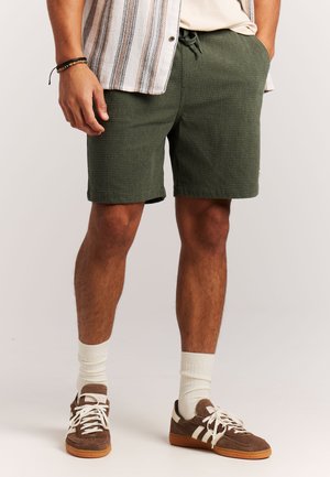 Man draagt olijfgroene, gestructureerde korte broek, beige overhemd, gestreept kort mouwen overhemd met knopen, witte sokken en bruine sneakers.