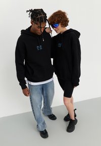 Black hoodies with front pockets and blue text detail, paired with light blue jeans and black sneakers. One person wears shorts, the other sunglasses.