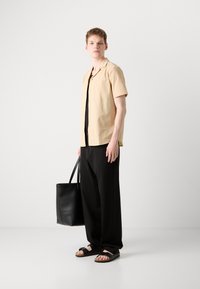 Hellbeige Kurzarmhemd, schwarzes Oberteil, locker sitzende schwarze Hosen und schwarze Tragetasche. Sandfarbene Sandalen mit Riemen. Einfaches Design.