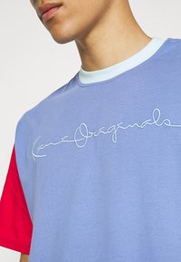 Triko s krátkým rukávem s levandulovým tělem, červenými rukávy a světle modrým límcem. Na přední straně je vyšito "Kari Originals" světle modrou kurzívou.