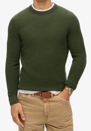 Mann trägt einen olivgrünen, strukturierten Pullover über einem weißen Hemd, beigen Hosen, braunem Gürtel und eine braune Armbanduhr mit Lederarmband.