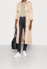 Beige trenchcoat med knappar, bärs över en ljusgrön topp och mörka jeans. Har vita sneakers och en svart handväska. Slät textur.