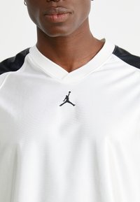 Jersey atlético blanco con acentos negros en los hombros, un cuello en V y un pequeño logo de Jumpman negro en el pecho, fabricado en tejido suave.