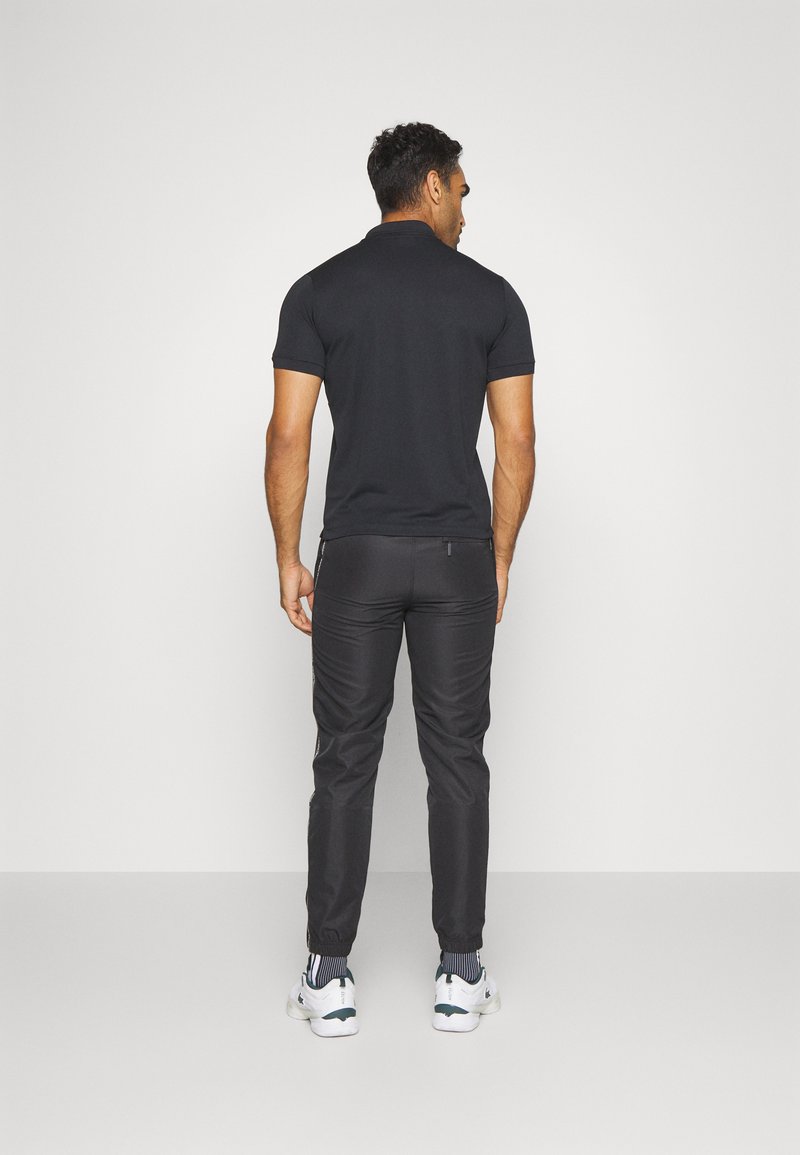 Polo noir à manches courtes, associé à un pantalon noir ajusté. Des chaussures blanches et noires présentent des accents rayés aux chaussettes.