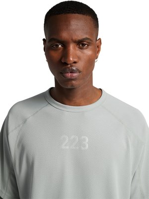 Mand med kort hår og øreringe iført en lysegrå tekstureret langærmet skjorte med nummeret "223" trykt på forsiden mod en hvid baggrund.