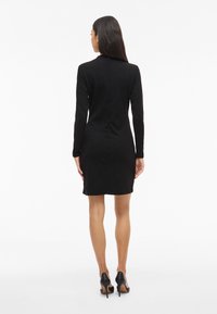 Femeie cu păr lung și închis la culoare, îmbrăcată într-o rochie neagră ajustată cu mâneci lungi și pantofi cu toc înalt negri, întoarsă cu spatele într-un fundal alb.