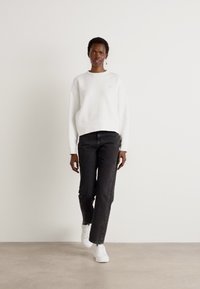 White sweatshirt with a crew neck, black jeans, and white sneakers. Model stands against a plain background, highlighting the outfit's colors.