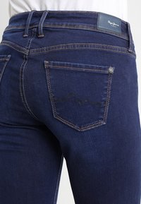 Jeans azul oscuro con un diseño ajustado, detalles de costura sutiles y un bolsillo trasero con marca que presenta un patrón distintivo.