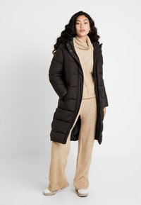 Long manteau en duvet noir avec capuche, présentant un design matelassé et des poches latérales. Porté par-dessus un pull en maille beige et un pantalon assorti.