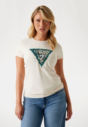 T-shirt crème à manches courtes présentant un graphique triangulaire vert orné de embellissements noirs et argentés ainsi que de texte. Jeans bleus.
