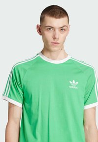 Zeleno bombažno majico s belimi detajli, kratkimi rokavi in tremi belimi črtami na ramenih. Ima majhen bel Adidas logotip na prsih.