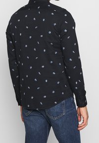 Maglietta nera a maniche lunghe con piccoli dettagli blu a motivo. Realizzata in tessuto liscio, presenta polsini con bottone e colletto standard.