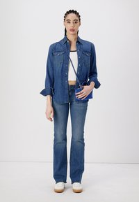 Donna con capelli intrecciati, indossa una camicia di denim e jeans, un crop top bianco, sneakers bianche, mentre tiene una piccola borsa a tracolla blu.