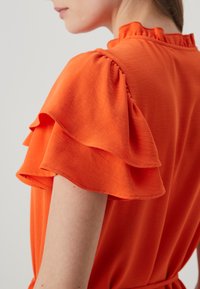 Πορτοκαλί μπλούζα κοντού μανικιού με στρωτές ρίγες, απαλή υφή και ψηλό λαιμό. Το ύφασμα κρέμεται απαλά στους ώμους.