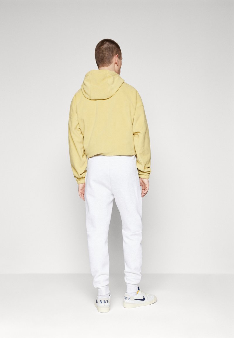 Persona vistiendo un sudadera con capucha de color amarillo claro y pantalones de chándal blancos, de espaldas, de pie sobre un fondo blanco liso.