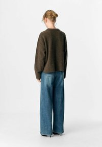 Frau mit blonden Haaren im Dutt, die einen dunkelbraunen Pullover und weit geschnittene blaue Jeans trägt, steht mit dem Rücken zu einem schlichten Hintergrund.