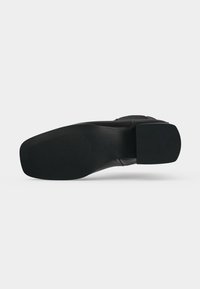Semelle de chaussure habillée noire avec zones de grip texturées et talon bas et large, vue de dessous sur un fond uni.