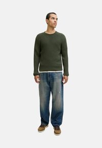 Mann in olivgrünem, grob gestricktem Pullover, lockeren blauen Jeans und braunen Schuhen, der vor einem schlichten hellen Hintergrund steht und nach rechts schaut.